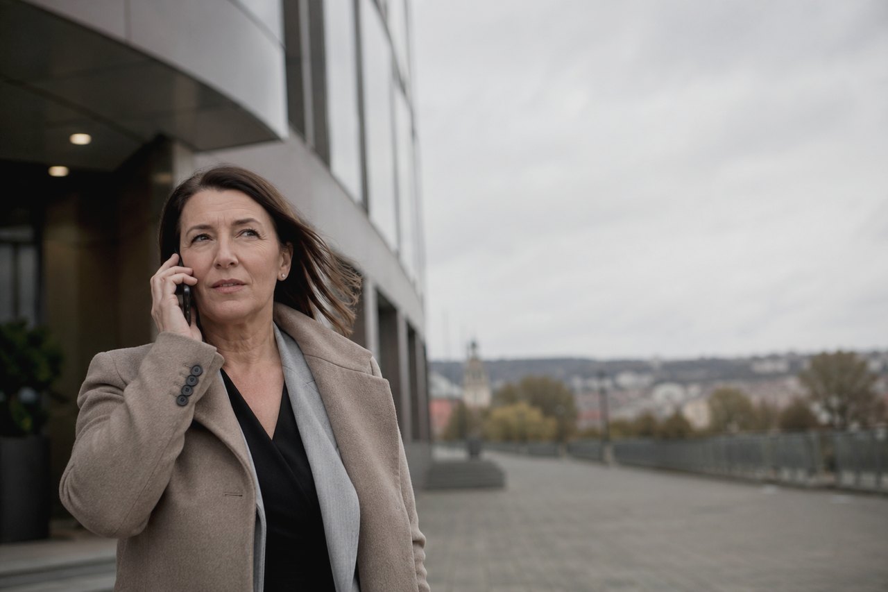 Podnikatelka v béžovém kabátě a šedé šále telefonuje před moderní pražskou kancelářskou budovou; v pozadí se pod zataženou podzimní oblohou rozprostírá panorama pražských střech s věží kostela.