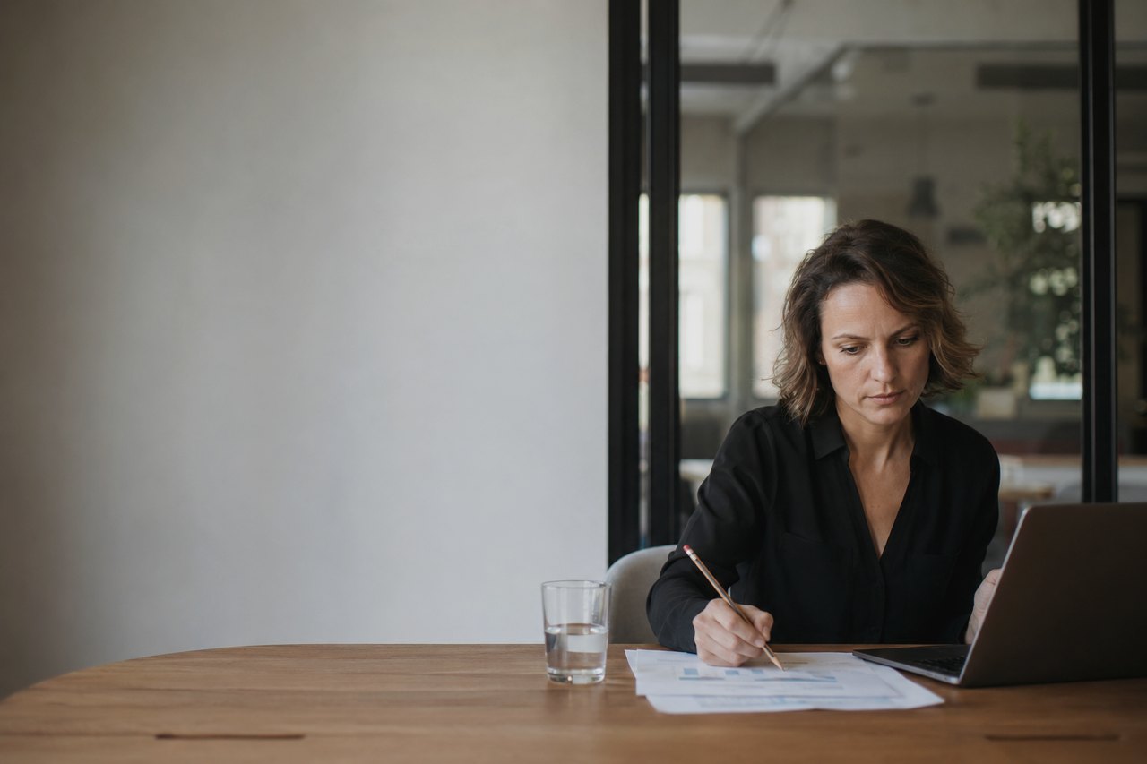 Žena v černé košili sedí u dřevěného stolu, tužkou si prochází vytištěné grafy a tabulky, vedle má otevřený notebook a sklenici vody; v pozadí prosklená stěna a světlá moderní kancelář.
